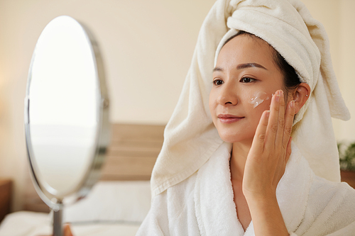Positive young woman in bathrobe looking at magnifying mirror and applying anti-aging lotion