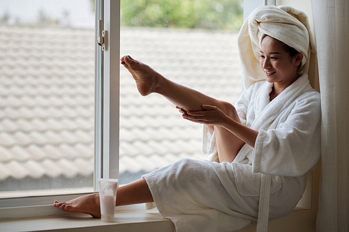 Cheerful young Asian woman sitting on window sill and applying calming aromatic body moisturizer on her legs