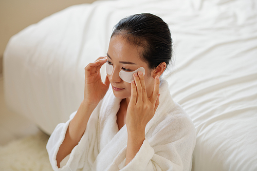 Pensive young Asian woman in bathrobe sitting on floor in bedroom and applying moisturizing patches