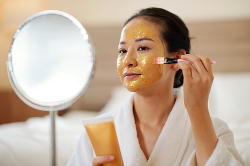 Smiling young woman applying soothing and nourishing golden gel mask on face after shower