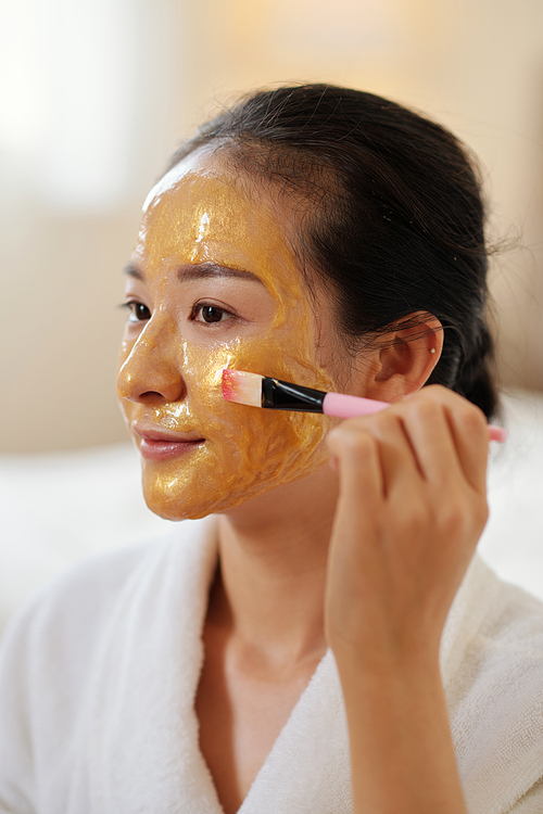 Positive young woman applying smoothing and softening face mask with particles