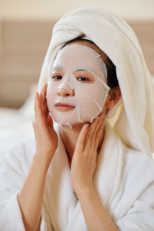 Portrait of positve young woman applying soothing and hydrating sheet mask on face after morning shower