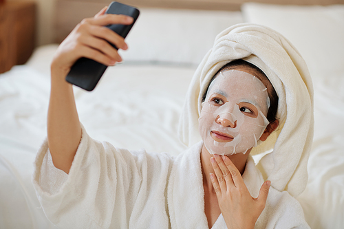 Young woman with pore tightening sheet mask on face taking selfie for blog