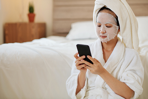 Serious young woman with sheet mask on face texting friends after morning shower