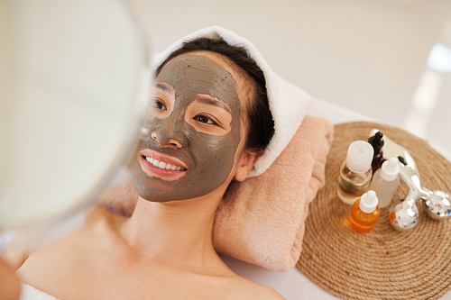 Smiling young Asian woman relaxing in spa salon with deep cleaning charcoal mask on her face