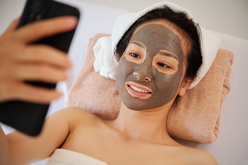 Smiling young woman with clay mask on face relaxing on bed in spa salon and video calling her friend