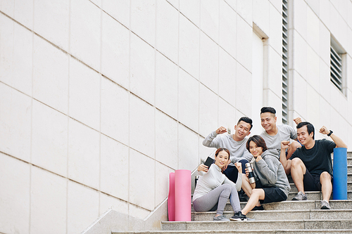 Group of excited young Asian people sitting on steps after sports training and posing for selfie and making fist pump gesture