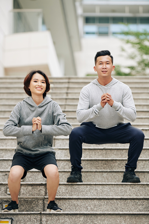 Cheerful young Vietnamese couple doing squats together after running on city streets in the morning