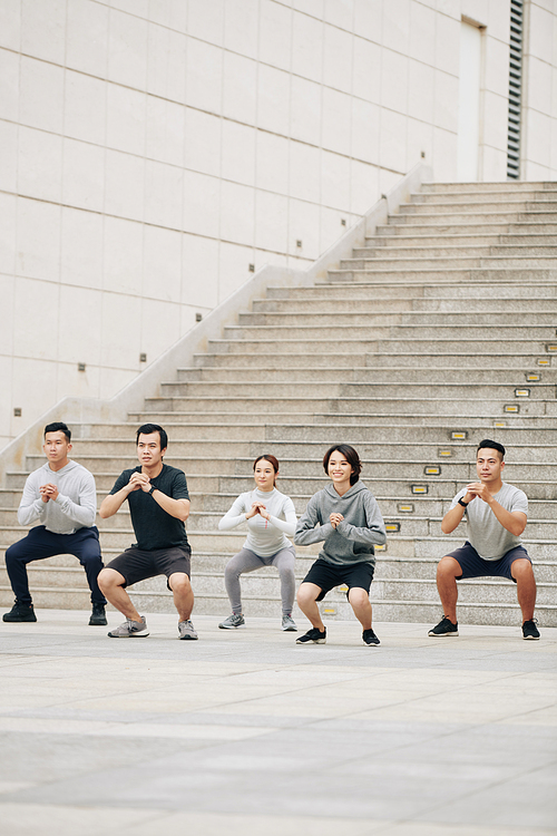 Cheerful young Vietnamese muscular athletic fit people doing squats together outdoors