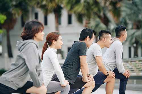 Young people doing deep lunges after morning jog in city park to stretch muscles and strengthen legs