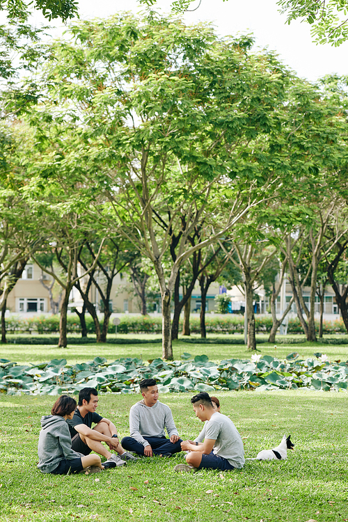 Group of young Asian people sitting on grass on university campus or in city park and discussing news