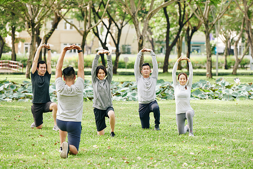 Group of young fit Vietnamese people following coach when doing exercise in city park