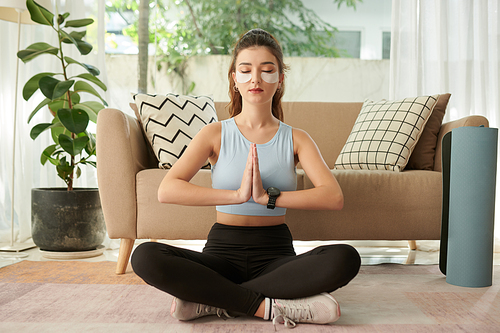 Young woman sitting in lotus position, keeping hands in namaste gesture when meditating at home