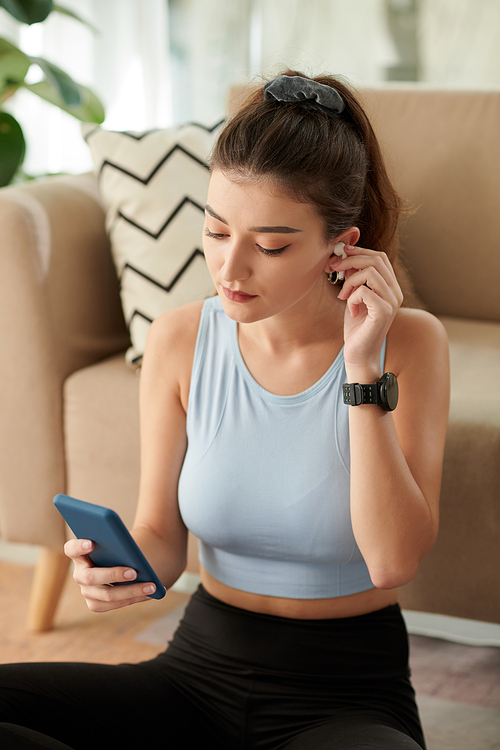 Young woman inserting earbud to listen to music on smartphone