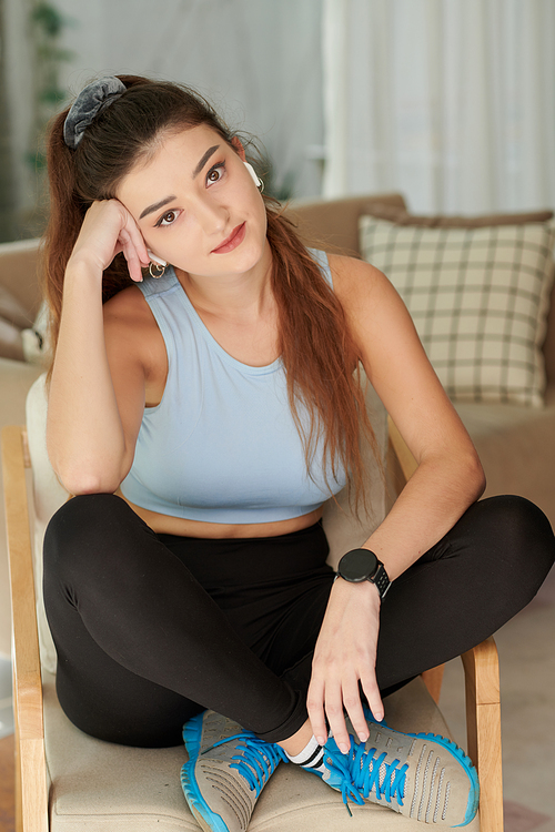 Dreamy smiling young woman in sportswear sitting on chair and smiling at camera