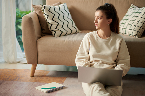 Serious young woman in loungewear sitting on floor at home and working on laptop