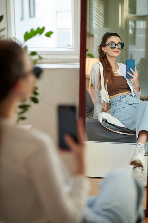 Young woman in sunglasses sitting on bed and taking selfie in mirror