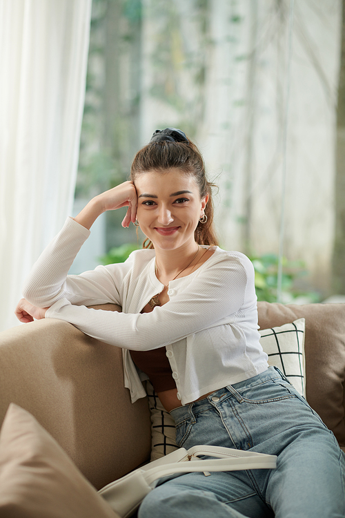 Lovely smiling young woman sitting on couch at home and looking at camera