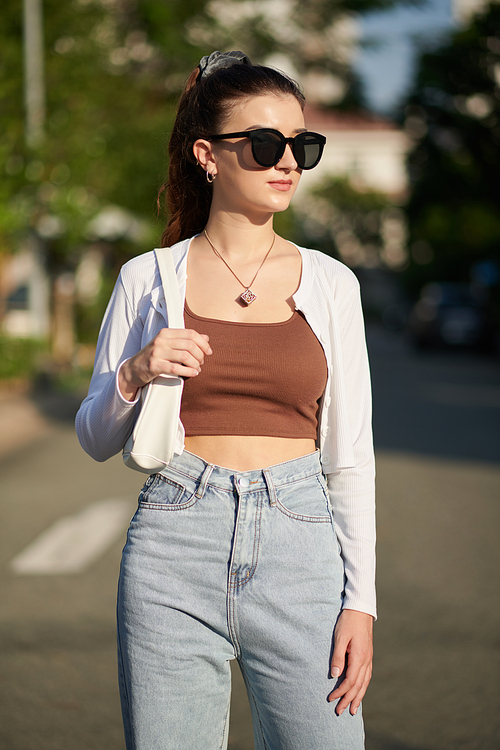 Portrait of young woman in high waisted jeans, brown top and short cardigan standing outdoors and looking away