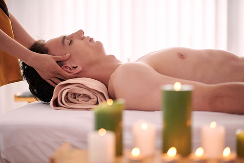 Side view of young serene and relaxed man lying on couch while masseuse standing behind and giving him professional massage in spa salon