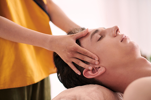 Hands of young masseuse on temples of serene multiethnic guy keeping head on rolled soft towel while relaxing in luxurious spa salon