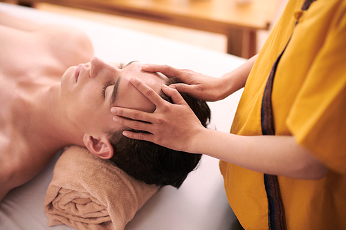 Young female physiotherapist in uniform keeping hands on forehead of male client during acupressure procedure or professional massage