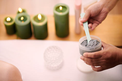 Closeup image of beautician mixing clay mask in plastic jar before applying it