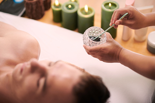 Hands of young female beautician mixing dry cosmetic clay with water in small jar while standing by relaxed male client lying on couch