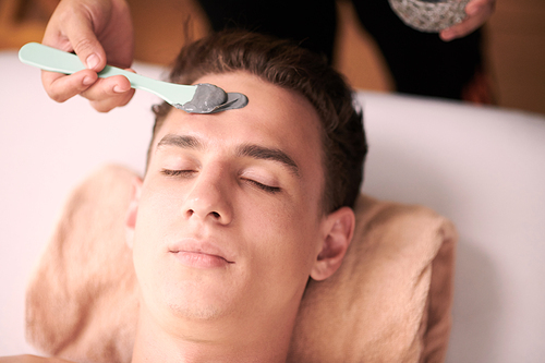 Above angle of relaxed and serene young man keeping head on rolled soft towel while beautician applying clay mask on his face in spa salon