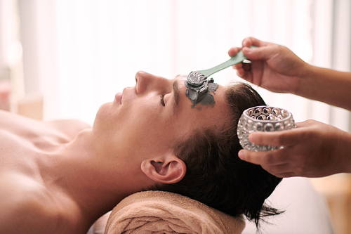 Side view of young serene man with closed eyes keeping head on rolled soft towel while female beautician applying clay mask on his face