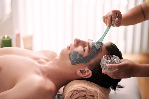 Young man getting purifying clay face treatment in beauty salon