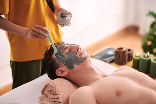 Young female beautician in yellow uniform standing by head of relaxed male client lying on couch and applying clay mask on his face