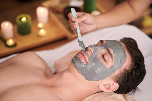 Beautician using plastic spatula when applying clay mask on face of young man