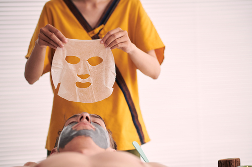 Young professional female beautician in yellow uniform holding sheet mask over serene male client with grey clay on face in spa salon