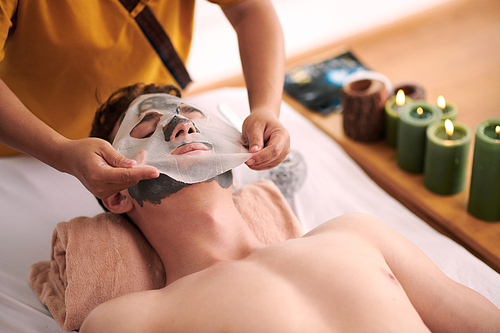 Beautician putting sheet mask on face of young man