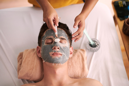 Hands of beautician massaging face of client with crystal roller after applying clay mask