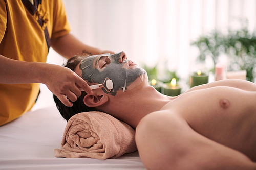 Side view of cosmetician hand with jade roller spreading sheet mask on face of young serene male client relaxing on couch in spa salon