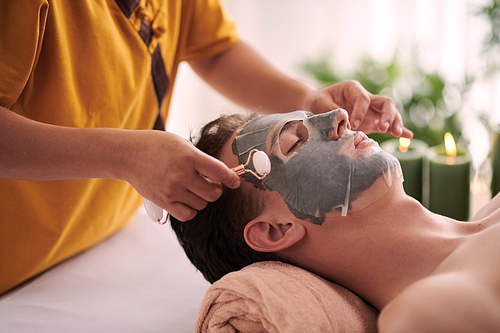 Cosmetologist giving rejuvenating face massage to man after applying face mask