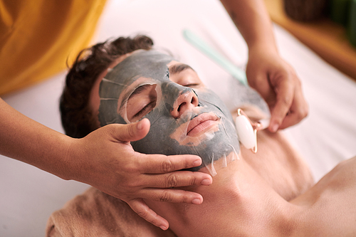 Hand of female beautician touching face of young male client with sheet mask over clay one and smoothing it with jade roller during procedure