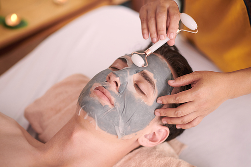 Young relaxed man with closed eyes getting facial massage with jade roller during procedure of hydrating and purifying skin in beauty salon