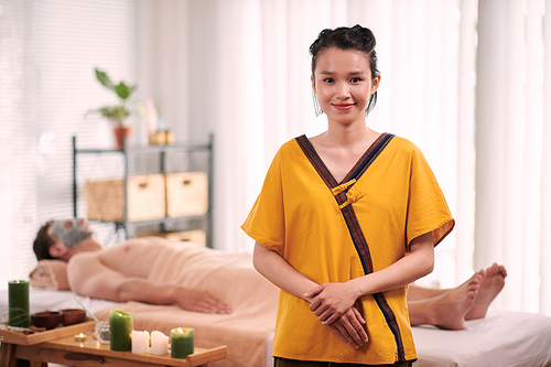 Pretty Asian girl in yellow uniform standing in front of camera against relaxed male client lying on massage couch during beautycare procedure