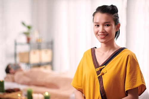 Young smiling Asian female beautician and masseuse in yellow uniform looking at you while standing in front of camera in luxurious spa salon