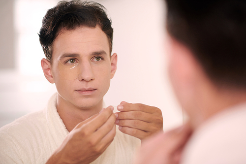 Portrait of young man applying undereye patches after morning shower