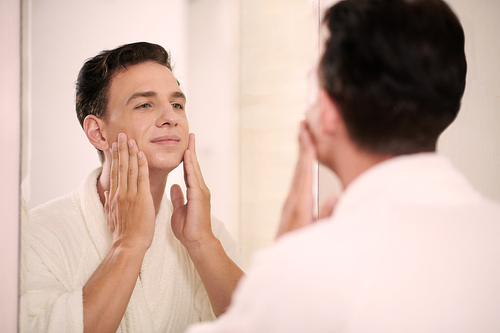 Young multiethnic man in white bathrobe touching his face while standing in front of mirror in bathroom and looking at his reflection