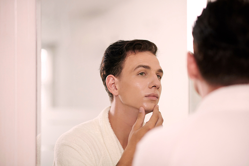 Young handsome man touching smooth and hydrated skin of his face and looking in mirror in bathroom after washing with new facial mousse