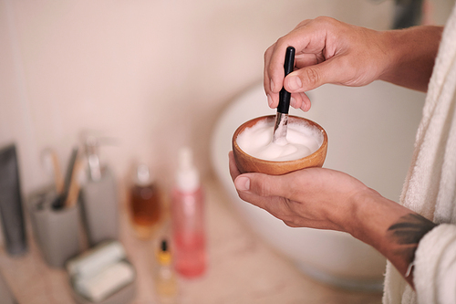 Hands of young man mixing soft foam with brush in small wooden bowl before procedure of shaving his face in bathroom in the morning