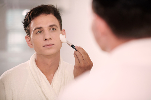 Young man in white soft bathrobe applying shaving foam on his face with brush while standing in front of mirror in bathroom in the morning