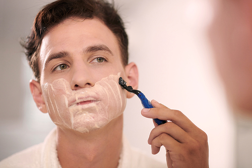 Young man shaving with razor while standing in front of mirror in bathroom and looking at his reflection during grooming procedure