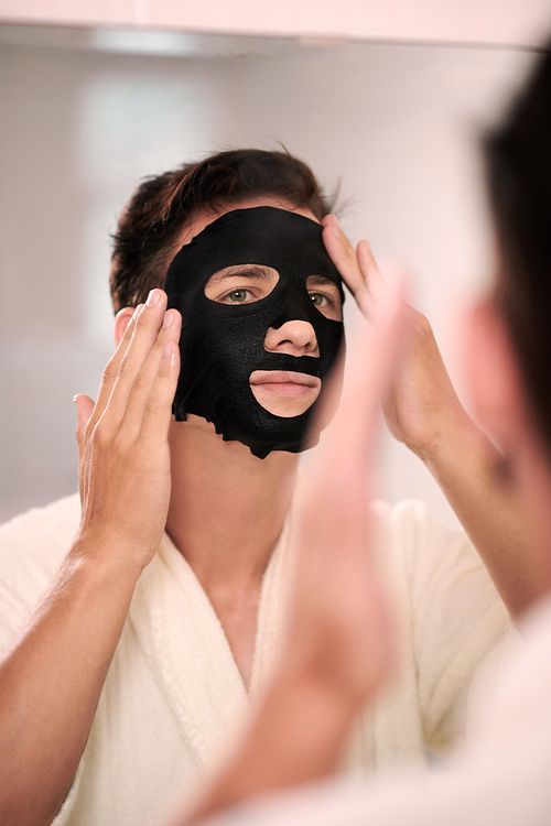 Young man applying purifying black sheet mask after morning shower