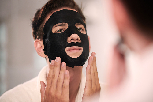 Young man with black hydrating sheet mask on his face standing in front of mirror in bathroom in the morning and looking at himself